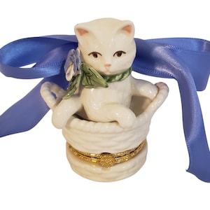 May include: A white ceramic cat figurine sits in a woven basket-shaped box, adorned with a blue ribbon. The cat wears a green scarf and is decorated with a small flower. The box has a gold-colored band around the middle.