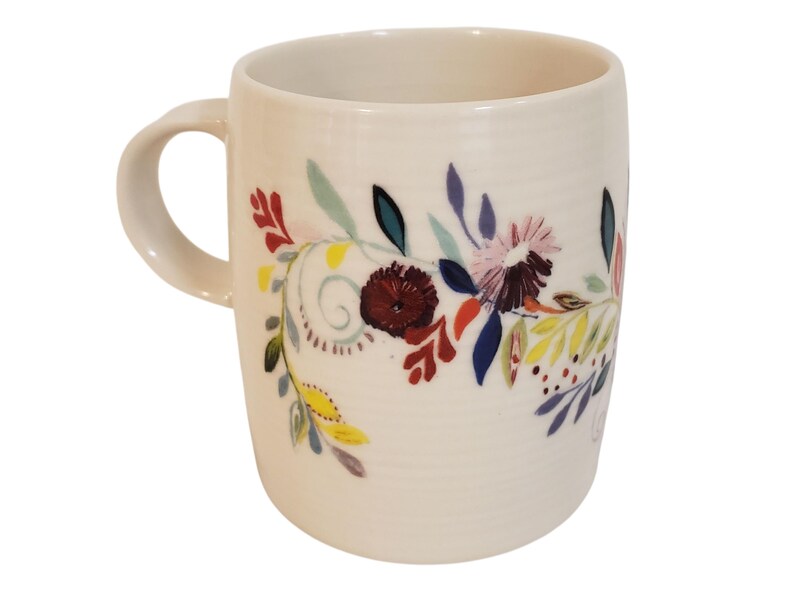 May include: Cream-colored ceramic mug with a curved handle. The mug is decorated with a floral design featuring burgundy, pink, blue, and yellow flowers and leaves. The design wraps around the mug's exterior.