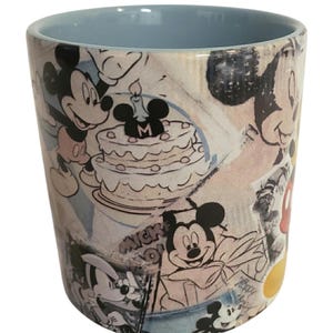 May include: A ceramic tumbler featuring a light blue interior and a wrap-around design with Mickey Mouse illustrations. The design includes images of Mickey Mouse, a birthday cake, and the word "Mickey".