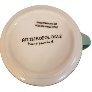 May include: A white ceramic mug with a light green handle and rim. The bottom of the mug features the text "ANTHROPOLOGIE hand painted" in black script. The mug is labeled "Dishwasher & Microwave Safe Made in China".