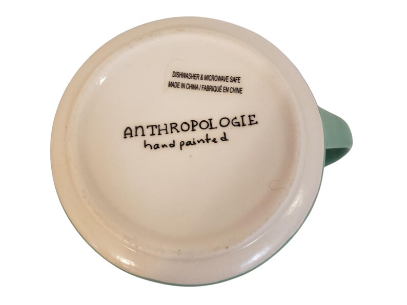 May include: A white ceramic mug with a light green handle and rim. The bottom of the mug features the text "ANTHROPOLOGIE hand painted" in black script. The mug is labeled "Dishwasher & Microwave Safe Made in China".