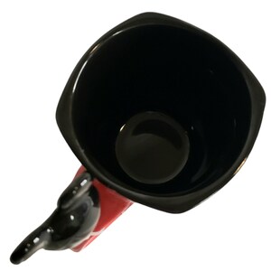 May include: A square-shaped ceramic mug with a glossy black interior and rim. The exterior features a vibrant red finish, and the handle is sculpted into the shape of a black character with rounded ears, extending outward from the side of the mug.