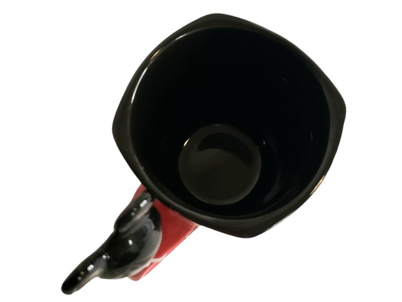 May include: A square-shaped ceramic mug with a glossy black interior and rim. The exterior features a vibrant red finish, and the handle is sculpted into the shape of a black character with rounded ears, extending outward from the side of the mug.