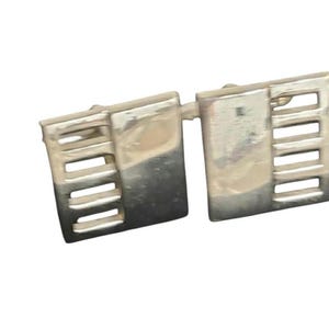 May include: Silver-toned cufflinks with a rectangular design. Each cufflink features a series of horizontal slots on one side, creating a geometric pattern. The cufflinks are connected by a small bar.