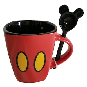 May include: A red ceramic mug with a black interior and two yellow oval shapes on the front. The mug features a matching black spoon with a handle shaped like two rounded ears, which rests in a slot integrated into the side of the mug handle.