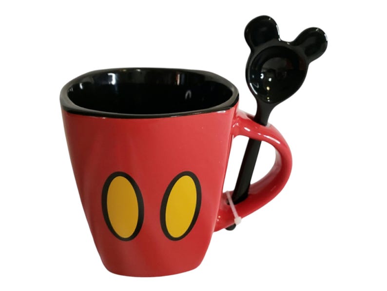 May include: A red ceramic mug with a black interior and two yellow oval shapes on the front. The mug features a matching black spoon with a handle shaped like two rounded ears, which rests in a slot integrated into the side of the mug handle.
