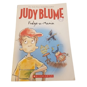 Puede incluir: Un libro de bolsillo titulado "Fudge-a-Mania" de Judy Blume. La portada presenta una ilustración de un niño con una camisa amarilla y una gorra roja, con otros personajes de dibujos animados y texto. El logotipo de Scholastic es visible.
