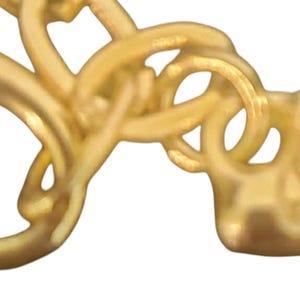 May include: Close-up of a gold-coloured jewellery clasp. The clasp features intertwined loops and a heart-shaped end. The metal has a polished finish, reflecting light. The design is intricate and detailed.