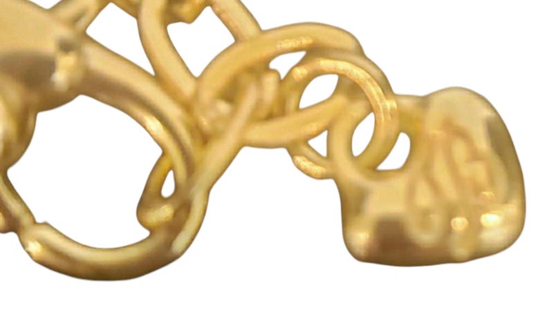 May include: Close-up of a gold-coloured jewellery clasp. The clasp features intertwined loops and a heart-shaped end. The metal has a polished finish, reflecting light. The design is intricate and detailed.