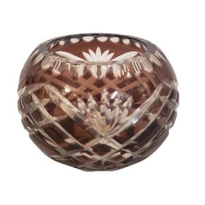 May include: A round, smoke-coloured glass bowl featuring intricate, hand-cut geometric patterns and floral motifs. The surface displays a combination of clear and frosted glass sections, creating a textured, multifaceted appearance with a wide opening.