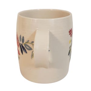May include: Cream-colored ceramic mug with a curved handle. The mug is decorated with floral designs in red, blue, and yellow. The mug is a standard size for coffee or tea.