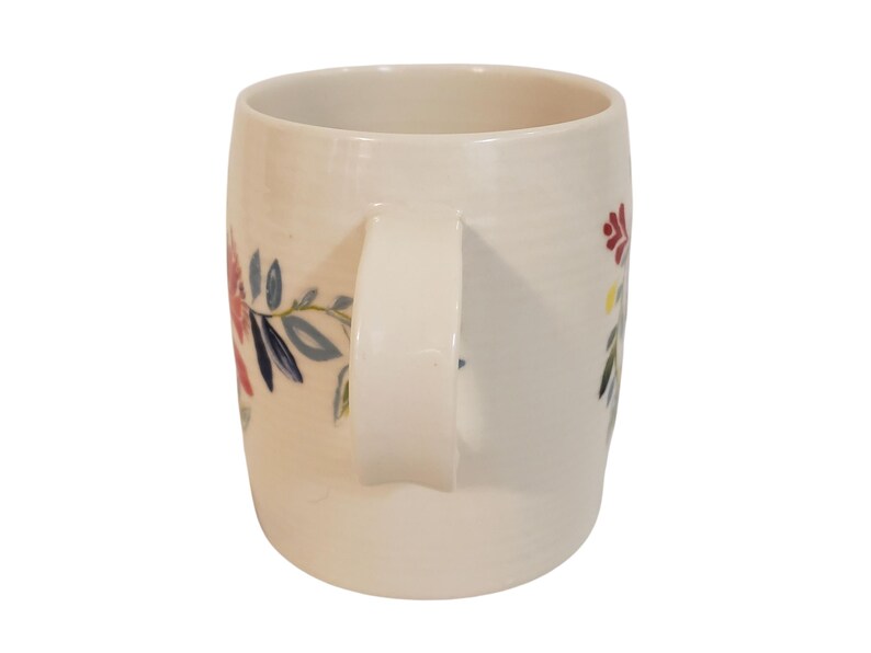 May include: Cream-colored ceramic mug with a curved handle. The mug is decorated with floral designs in red, blue, and yellow. The mug is a standard size for coffee or tea.