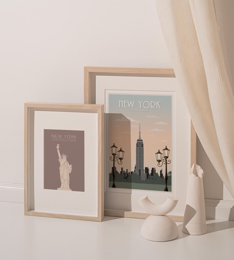May include: Two framed prints of New York City landmarks. The left print features a brown background with a line drawing of the Statue of Liberty. The right print features a skyline with the Empire State Building and streetlights. Both prints have a light wood frame.