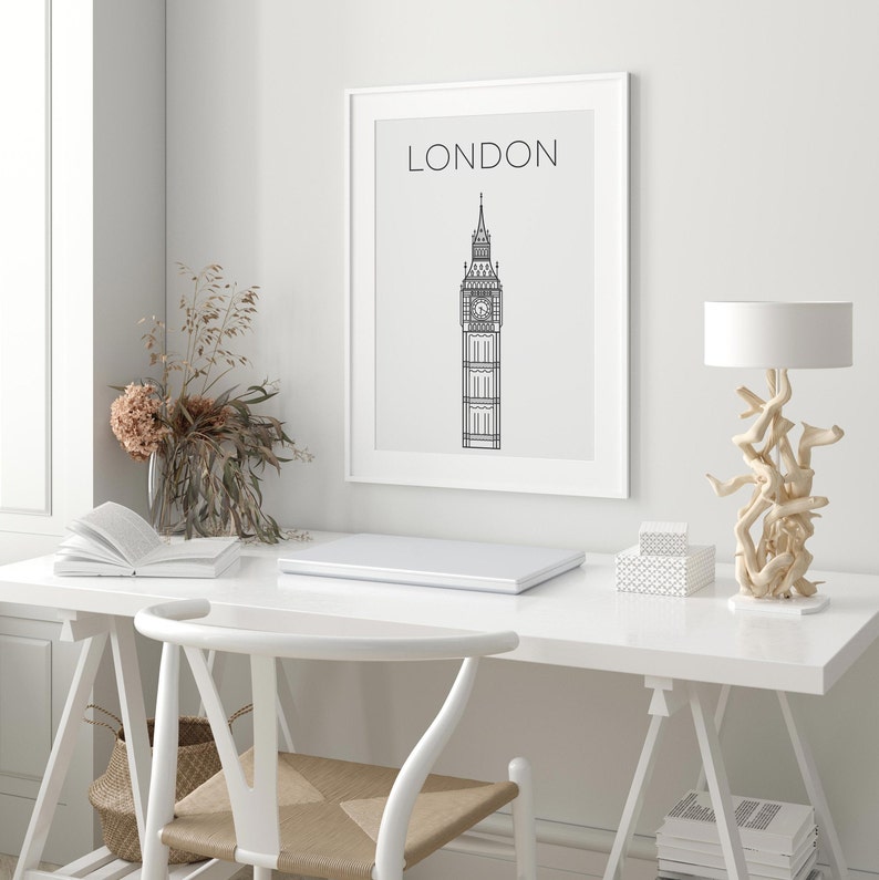 May include: A white framed print with a black line drawing of Big Ben and the text "LONDON" above it. The print is hanging on a white wall above a white desk with a laptop, a book, a lamp, and a chair.