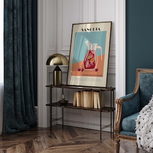 May include: A framed art print featuring a Sangria illustration with a pitcher, fruit, and the text "Sangria" and "Originates in Spain." The artwork is displayed on a dark brown shelf with a gold lamp and books. The room has teal curtains and a blue armchair.