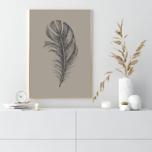 May include: A black and white illustration of a feather on a beige background. The illustration is framed in a gold frame and is hanging on a white wall.