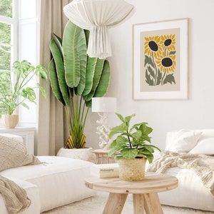 May include: A living room interior with a white couch, a coffee table, a potted plant, and a framed print of stylized sunflowers. The print has a beige background with green leaves and yellow sunflowers.