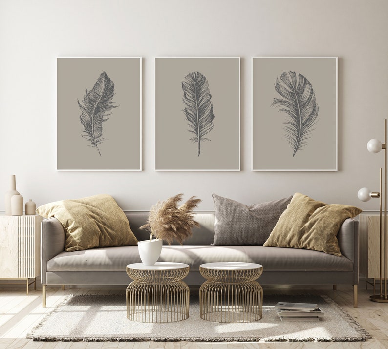 May include: Three framed prints of black and white feather illustrations on a beige background. The prints are hanging on a white wall above a gray sofa with brown throw pillows.