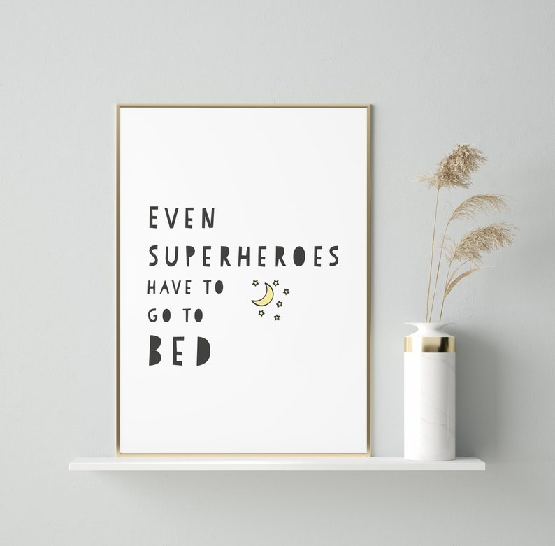 Puede incluir: Un p&oacute;ster blanco con texto negro que dice "Incluso los superh&eacute;roes tienen que irse a la cama" con una luna creciente amarilla y estrellas. El p&oacute;ster est&aacute; enmarcado en oro y est&aacute; en un estante blanco.