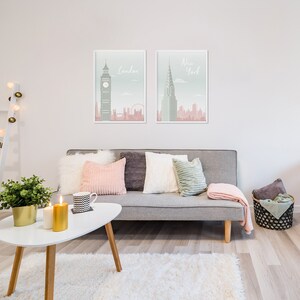 May include: Two framed art prints of London and New York City hang above a gray sofa with decorative pillows and a pink throw blanket. A white coffee table holds candles and a plant. The room has a modern, minimalist aesthetic.