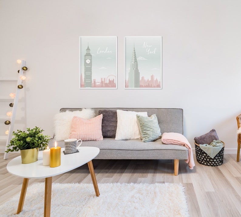 May include: Two framed art prints of London and New York City hang above a gray sofa with decorative pillows and a pink throw blanket. A white coffee table holds candles and a plant. The room has a modern, minimalist aesthetic.