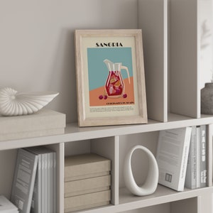 May include: A framed print featuring a Sangria illustration with a pitcher filled with fruit and the text "Sangria" at the top. The print is displayed on a white shelf with decorative items, including a seashell, boxes, and books. The color palette includes beige, red, and teal.