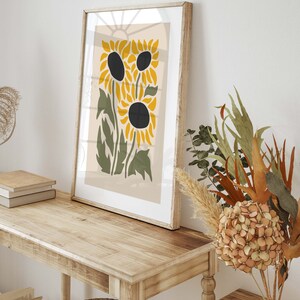 May include: A framed print of a stylized sunflower design with yellow petals and black centers. The print is on a beige background and is framed in a light wood frame.