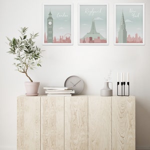 May include: Three framed art prints featuring cityscapes of London, Reykjavik, and New York. Each print has a white frame and a pastel color scheme with the city name in a script font. The prints are displayed above a light wood cabinet.