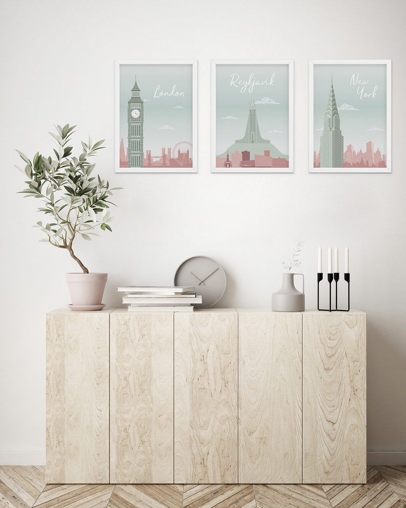 May include: Three framed art prints featuring cityscapes of London, Reykjavik, and New York. Each print has a white frame and a pastel color scheme with the city name in a script font. The prints are displayed above a light wood cabinet.
