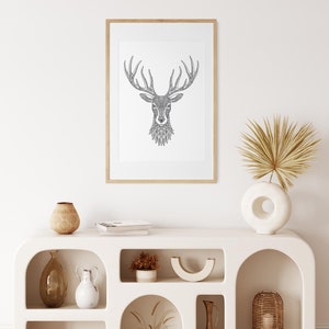 May include: A framed black and white line drawing of a deer head with intricate details. The frame is light wood and the drawing is on a white background. The drawing is hanging on a white wall above a white shelf with decorative items.