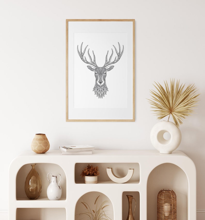 May include: A framed black and white line drawing of a deer head with intricate details. The frame is light wood and the drawing is on a white background. The drawing is hanging on a white wall above a white shelf with decorative items.