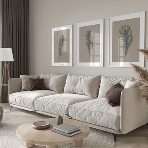 May include: A white sectional sofa with brown throw pillows sits in a living room with a light wood floor. Three framed prints with black and white drawings of feathers hang on the wall behind the sofa. A dried grass plant in a brown vase sits on the floor to the right of the sofa.