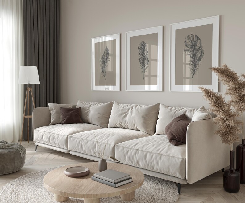 May include: A white sectional sofa with brown throw pillows sits in a living room with a light wood floor. Three framed prints with black and white drawings of feathers hang on the wall behind the sofa. A dried grass plant in a brown vase sits on the floor to the right of the sofa.
