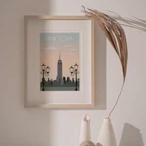 Puede incluir: Una impresi&oacute;n enmarcada de un paisaje urbano con el Empire State Building en el centro. La impresi&oacute;n est&aacute; en un marco de madera claro y tiene un fondo azul claro con el texto "NEW YORK" en la parte superior. La impresi&oacute;n est&aacute; colgada en una pared blanca con una palmera seca y dos jarrones de cer&aacute;mica blanca en primer plano.