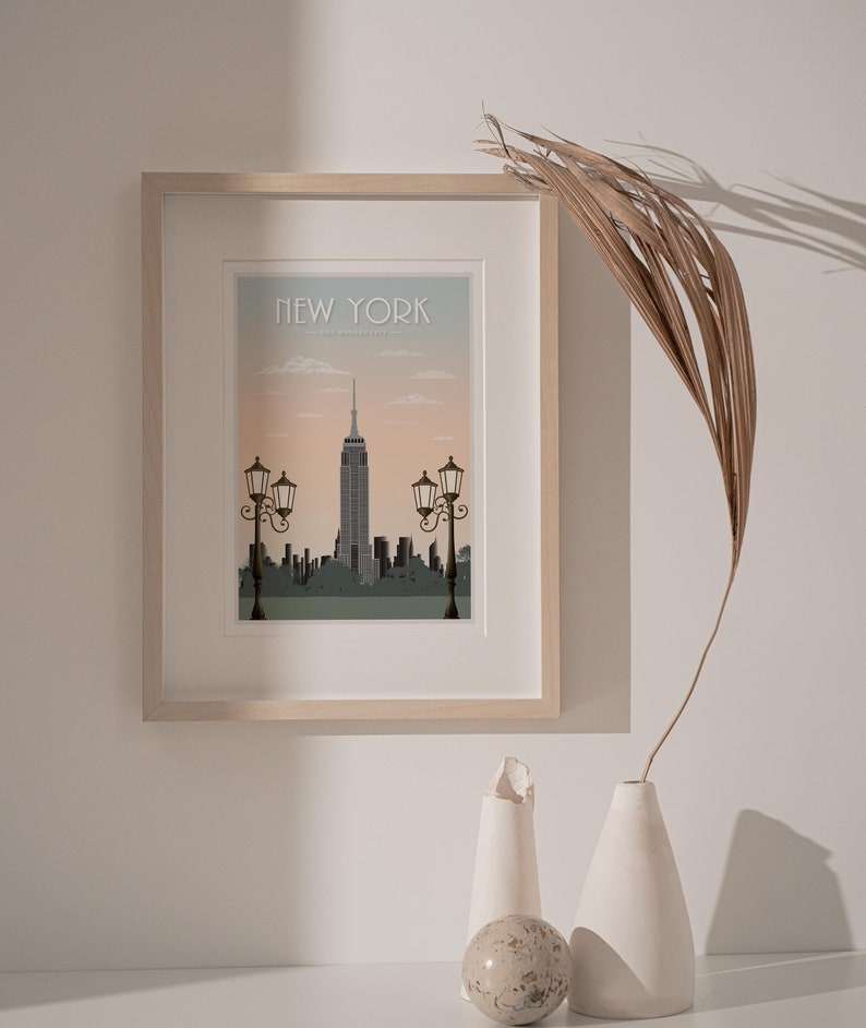 Puede incluir: Una impresi&oacute;n enmarcada de un paisaje urbano con el Empire State Building en el centro. La impresi&oacute;n est&aacute; en un marco de madera claro y tiene un fondo azul claro con el texto "NEW YORK" en la parte superior. La impresi&oacute;n est&aacute; colgada en una pared blanca con una palmera seca y dos jarrones de cer&aacute;mica blanca en primer plano.