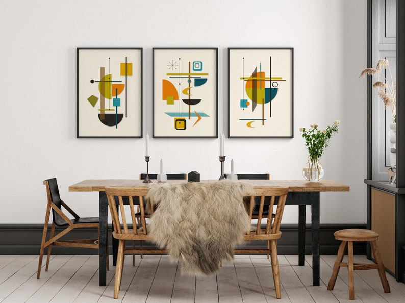 Puede incluir: Tres impresiones de arte abstracto enmarcadas con formas geom&eacute;tricas en tonos de amarillo, naranja, azul, verde y marr&oacute;n. Las impresiones est&aacute;n colgadas en una pared blanca sobre una mesa de comedor de madera con dos sillas y una manta de piel.