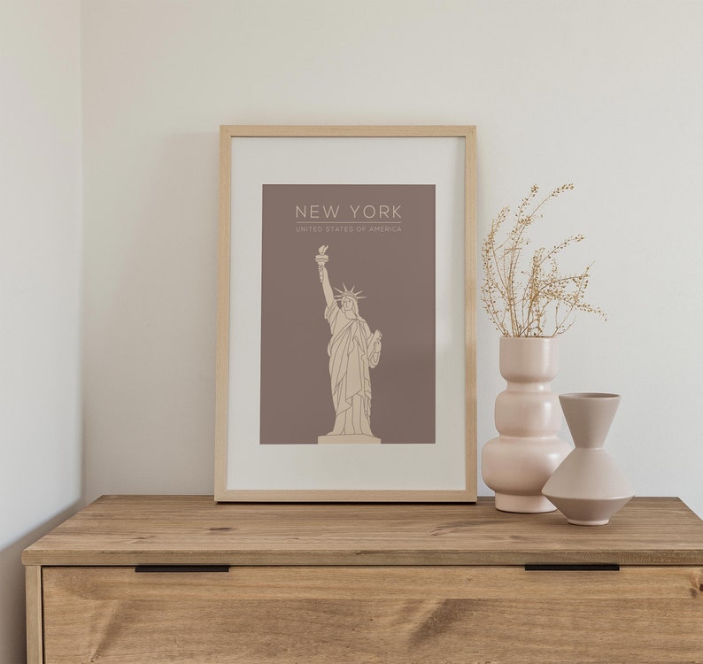 May include: A framed print with a brown background and a line drawing of the Statue of Liberty. The text "NEW YORK" and "UNITED STATES OF AMERICA" is printed in beige below the image.