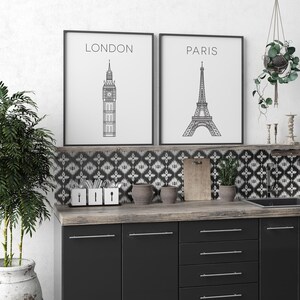 May include: Two black framed prints with white backgrounds and black line drawings of the Eiffel Tower and Big Ben. The prints are hanging on a white wall above a black and white patterned tile backsplash. The backsplash is above a dark wood countertop with black cabinets.