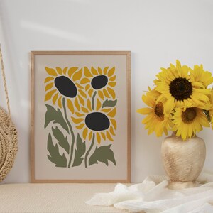 May include: A framed print of a stylized sunflower design in yellow, green, and brown. The print is on a cream background and is framed in a light wood frame.