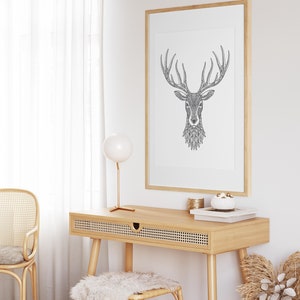 May include: A black and white line drawing of a deer head with antlers, framed in a light wood frame, hangs above a light wood desk with a woven rattan chair and a white fluffy stool. A gold table lamp with a white globe sits on the desk.