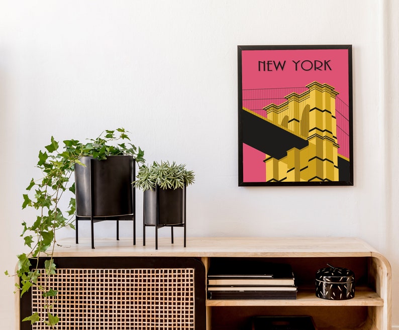 Puede incluir: Una impresi&oacute;n enmarcada de un puente amarillo sobre un fondo rosa con el texto "NEW YORK" en la parte superior. La impresi&oacute;n est&aacute; colgada en una pared blanca sobre un estante de madera con plantas y objetos decorativos.