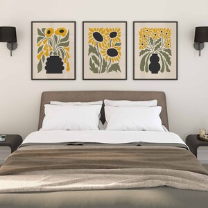 May include: Three framed prints of stylized floral arrangements with black silhouettes of sunflowers and other flowers in yellow and green hues. The prints are hanging on a white wall above a brown headboard with white bedding.