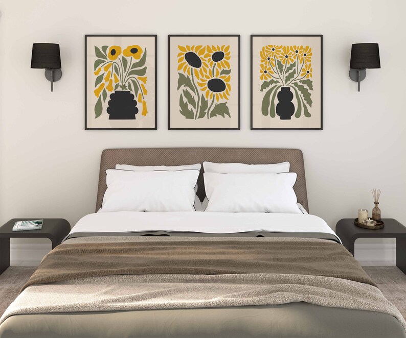 May include: Three framed prints of stylized floral arrangements with black silhouettes of sunflowers and other flowers in yellow and green hues. The prints are hanging on a white wall above a brown headboard with white bedding.