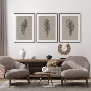 May include: Three framed prints of black and white line drawings of feathers on a beige background. The prints are hanging on a white wall above a wooden shelf with a vase, a bowl, and a decorative object. Two armchairs are in front of the shelf.