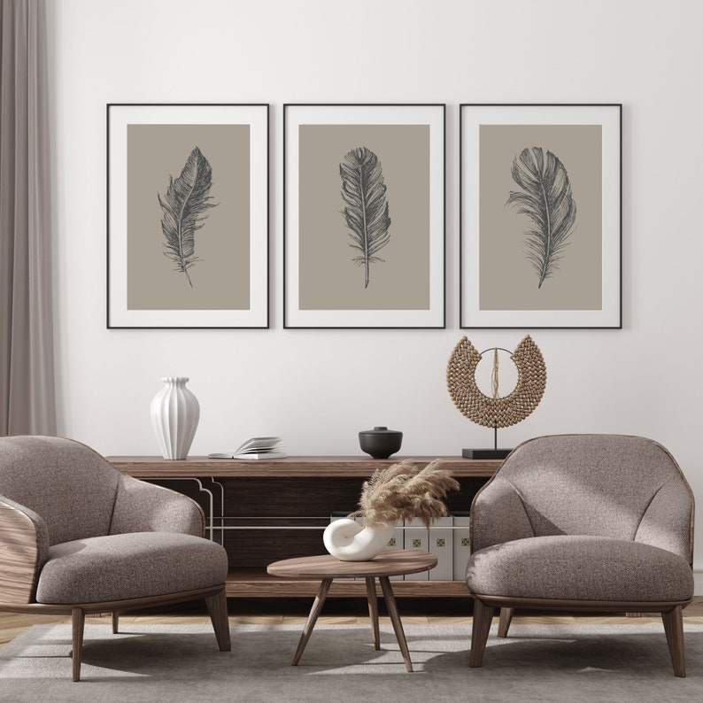 May include: Three framed prints of black and white line drawings of feathers on a beige background. The prints are hanging on a white wall above a wooden shelf with a vase, a bowl, and a decorative object. Two armchairs are in front of the shelf.