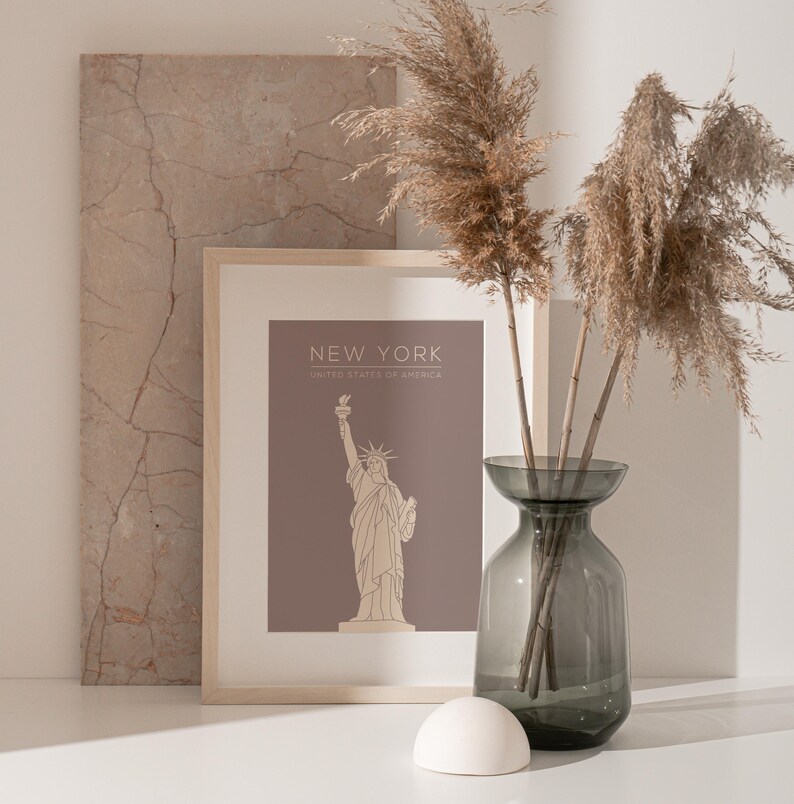 May include: A framed print with a brown background and a line drawing of the Statue of Liberty. The text "NEW YORK" and "UNITED STATES OF AMERICA" is printed below the image. The print is sitting on a white surface with a vase of dried pampas grass.