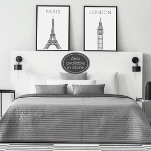 May include: Two black framed prints with white backgrounds. The print on the left features a line drawing of the Eiffel Tower with the word "PARIS" above it. The print on the right features a line drawing of Big Ben with the word "LONDON" above it. The prints are hanging on a white wall above a grey bed with white pillows and a grey bedspread.