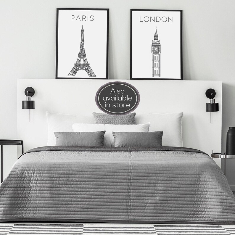 May include: Two black framed prints with white backgrounds. The print on the left features a line drawing of the Eiffel Tower with the word "PARIS" above it. The print on the right features a line drawing of Big Ben with the word "LONDON" above it. The prints are hanging on a white wall above a grey bed with white pillows and a grey bedspread.