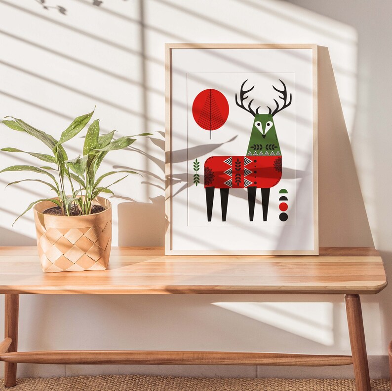 May include: A framed print of a stylized deer wearing a red and green sweater with geometric patterns. The deer is standing on a white background with a red circle and a green circle behind it.