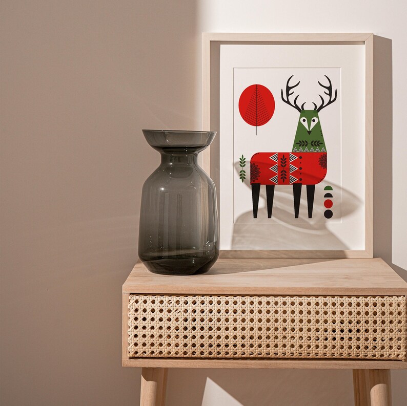 May include: A dark gray glass vase sits on a light wood table with a woven rattan drawer. Behind the vase is a framed print of a stylized deer wearing a red and green sweater.